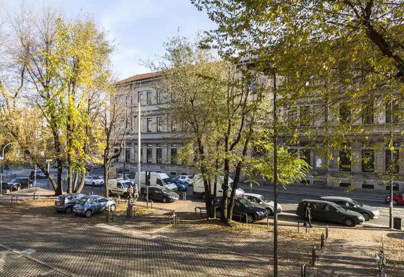 Vista esterna verso la strada con un parcheggio lastricato, auto parcheggiate e alberi maturi, di fronte a una facciata classica. Luce autunnale brillante e buona visibilità rendono la scena esterna invitante.
