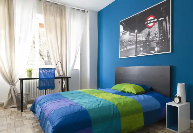 Bright, well-staged bedroom with a double bed framed against a deep blue accent wall, colorful bedding and a small desk by the window that brings abundant natural light.