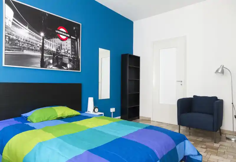 Bright, modern bedroom with a colorful double bed, a comfortable armchair and minimalist shelving — ideal for short-term rentals.