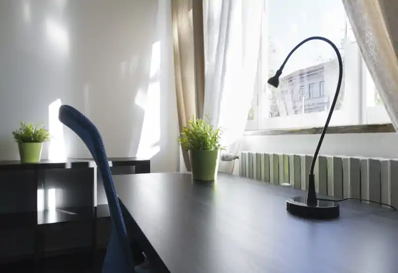 Angolo soggiorno/studio luminoso e minimale con una scrivania ordinata vicino alla finestra, una lampada da lettura e piante in vaso che creano un piacevole spazio di lavoro.