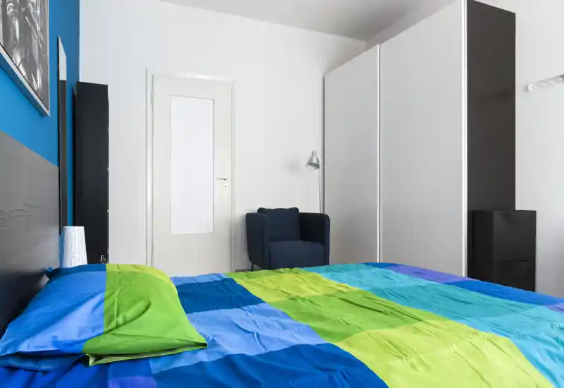 Bright, modern bedroom with a large bed dressed in colorful blue and green linens, a comfortable armchair and a spacious white wardrobe — ideal for a fresh, tidy rental listing.