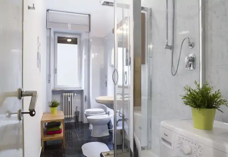 Bagno luminoso e ordinato con cabina doccia, WC, bidet e lavabo; spazio compatto con lavatrice e piante decorative che donano un tocco fresco.