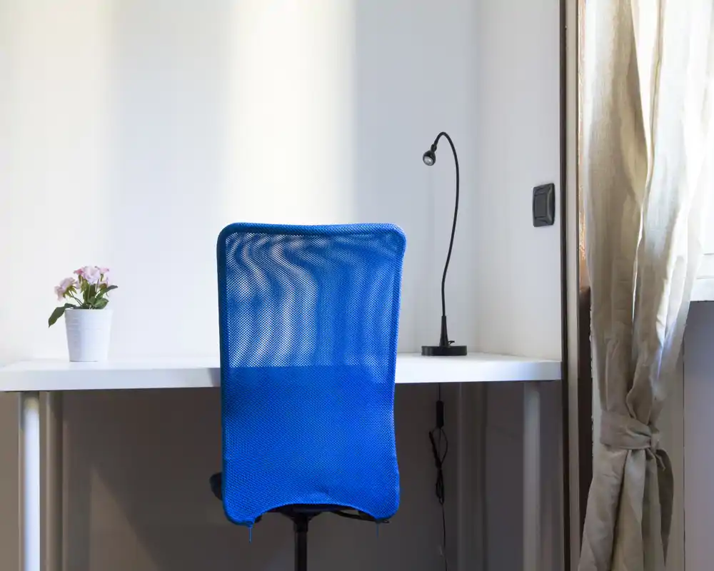 Rincón de trabajo en casa ordenado con silla azul de malla, escritorio blanco, lámpara pequeña y una planta junto a una ventana con cortina — luminoso, minimalista y funcional.