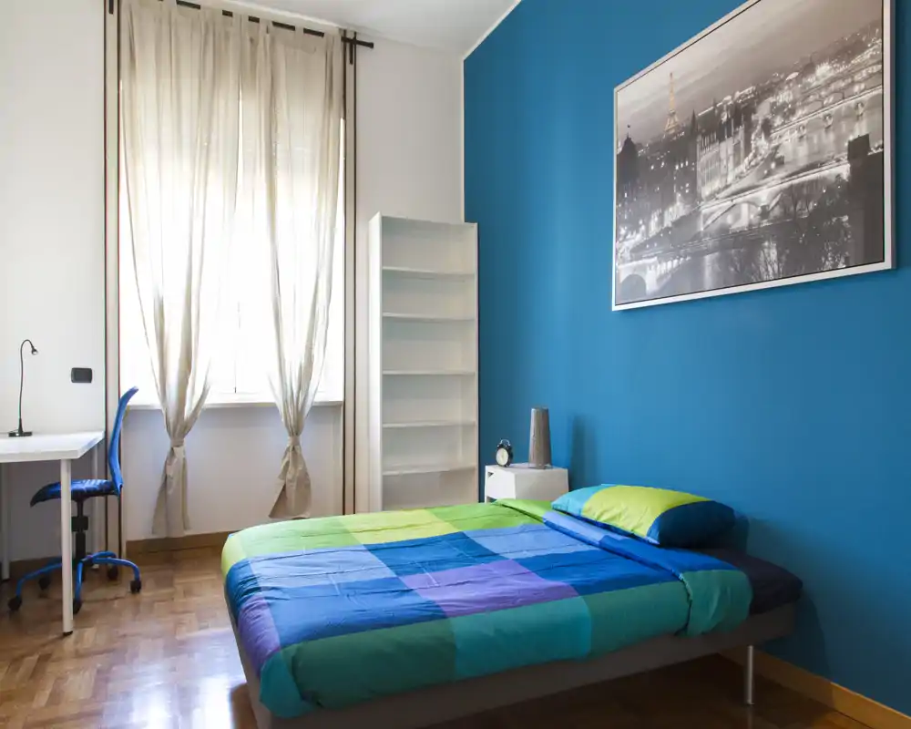 Dormitorio tranquilo y luminoso con cama doble frente a una pared azul vibrante, suelo de parquet, mesita y escritorio junto a la ventana — ideal para estancias cómodas o estudiantes.
