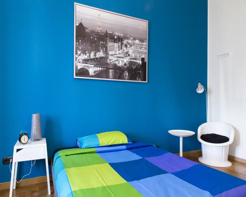 Dormitorio moderno y luminoso con una cama colorida frente a una pared azul intensa, mesita y sillón de lectura—estiloso y bien iluminado.