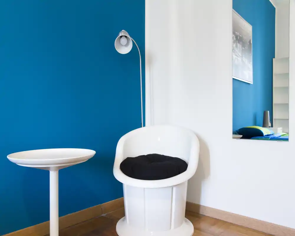 Rincón de sala luminoso y moderno con butaca blanca, mesita y lámpara de pie frente a una pared azul intensa — un asiento sencillo y elegante ideal para leer.