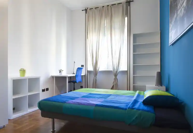 Camera da letto luminosa e moderna con letto matrimoniale con biancheria colorata, scrivania vicino alla finestra e scaffalatura minimal — ideale per studenti o professionisti in cerca di una stanza ordinata e ben illuminata.