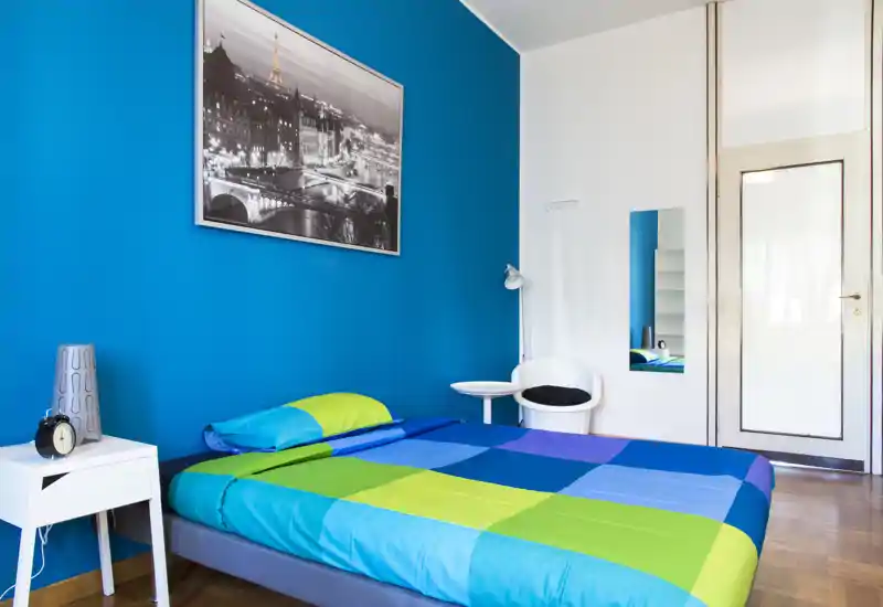 Camera da letto moderna e luminosa con letto matrimoniale colorato davanti a una parete blu intensa, comodino e lampada — pulita e accogliente.