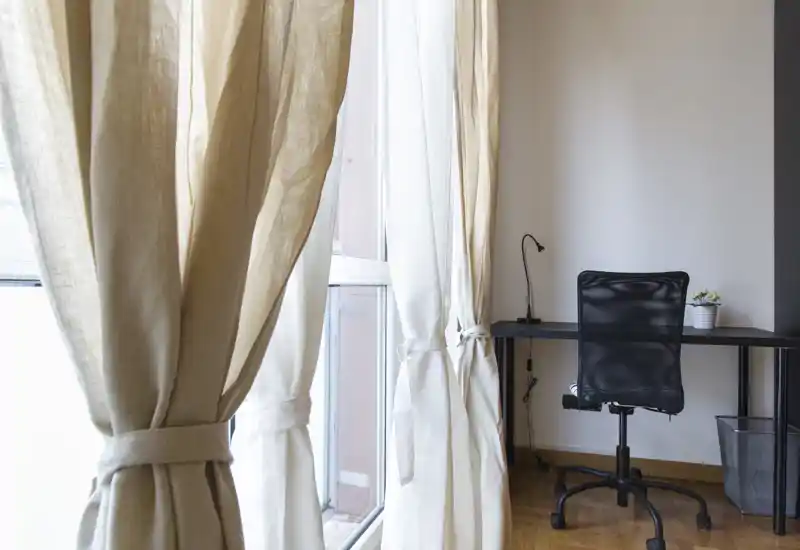 Angolo luminoso living/lavoro con ampie finestre e tende neutre raccolte, scrivania minimal e sedia ergonomica — perfetto come area di lavoro tranquilla con molta luce naturale.