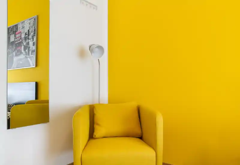 Angolo luminoso e accogliente con una poltrona imbottita gialla e cuscino coordinato su una parete gialla intensa, con lampada da terra e specchio che aggiungono profondità.