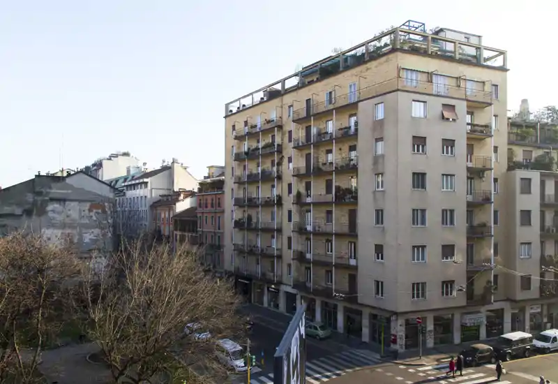 Veduta stradale di un edificio residenziale e del quartiere circostante, con balconi, negozi al piano terra e una piccola piazzetta alberata.