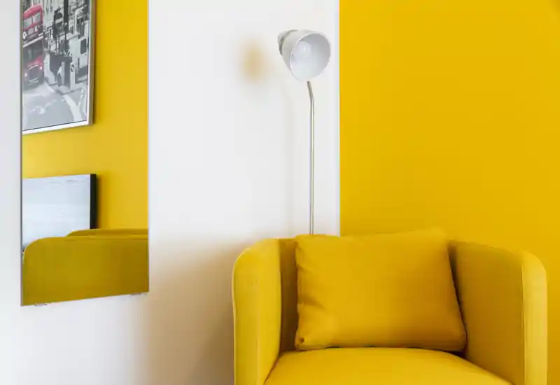 Angolo luminoso e moderno con una poltrona imbottita giallo senape, cuscino abbinato e lampada da terra su pareti bianche e gialle — un accogliente angolo salotto.