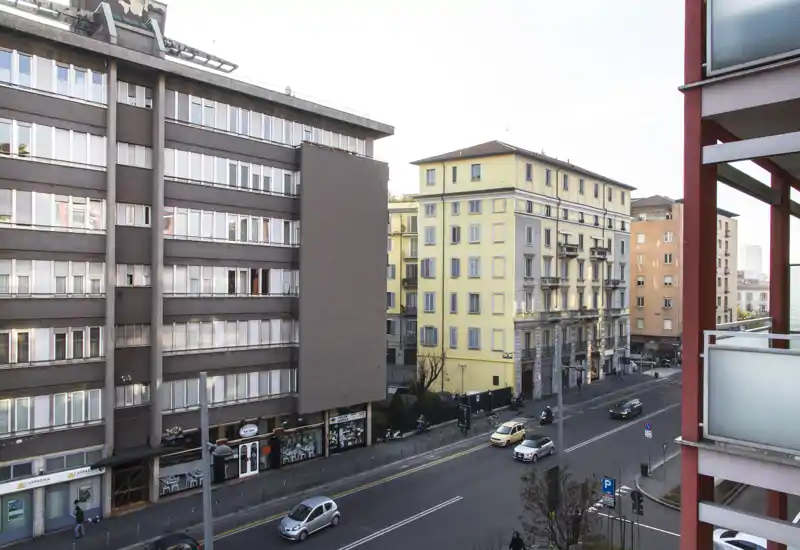 Vista dalla balconata su una strada urbana trafficata con edifici residenziali e auto in transito.