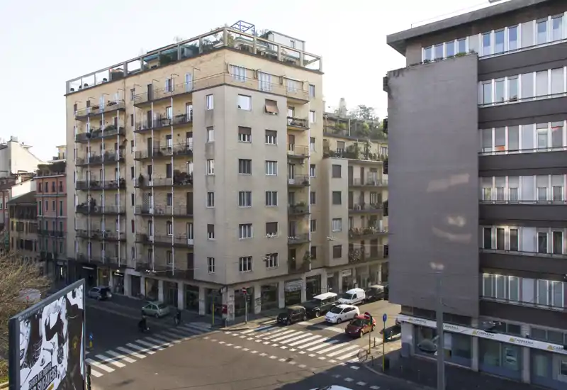 Street-level exterior view of an urban corner with multi-storey apartment buildings, balconies and a crosswalk — useful for showing location and façade condition.