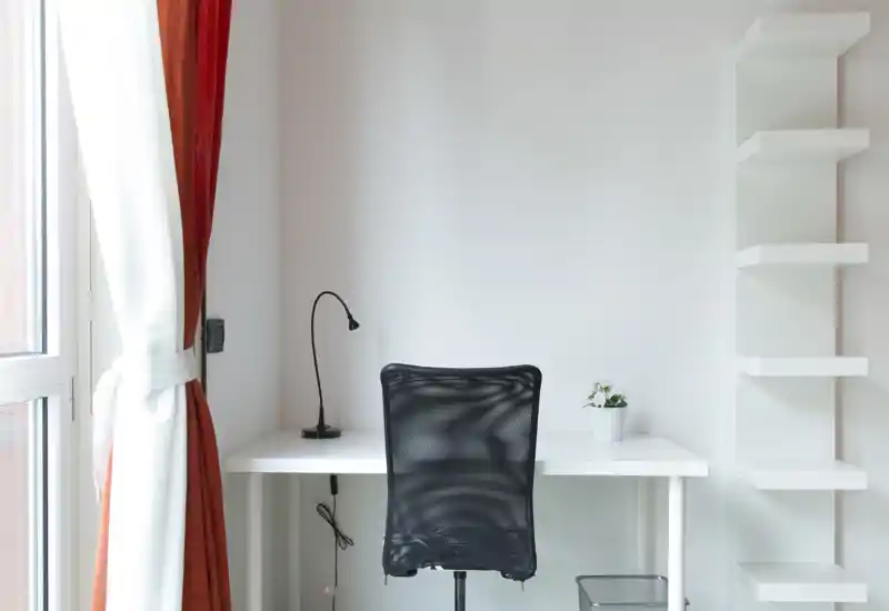 Bright, minimalist workspace with a white desk, black mesh office chair, small desk lamp and shelving — ideal for a focused work or study corner with natural light.
