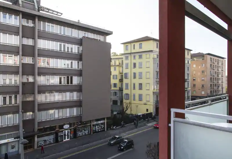 Balcony view overlooking a city street with nearby apartment buildings; glass and metal railing in the foreground and clear urban surroundings.