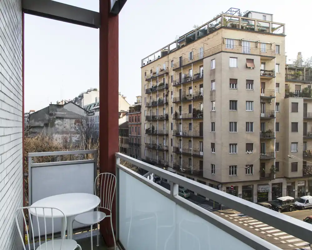 Balcone cittadino soleggiato con un piccolo tavolo rotondo e due sedie in metallo, affacciato su una strada e palazzi adiacenti — ideale per un caffè mattutino.