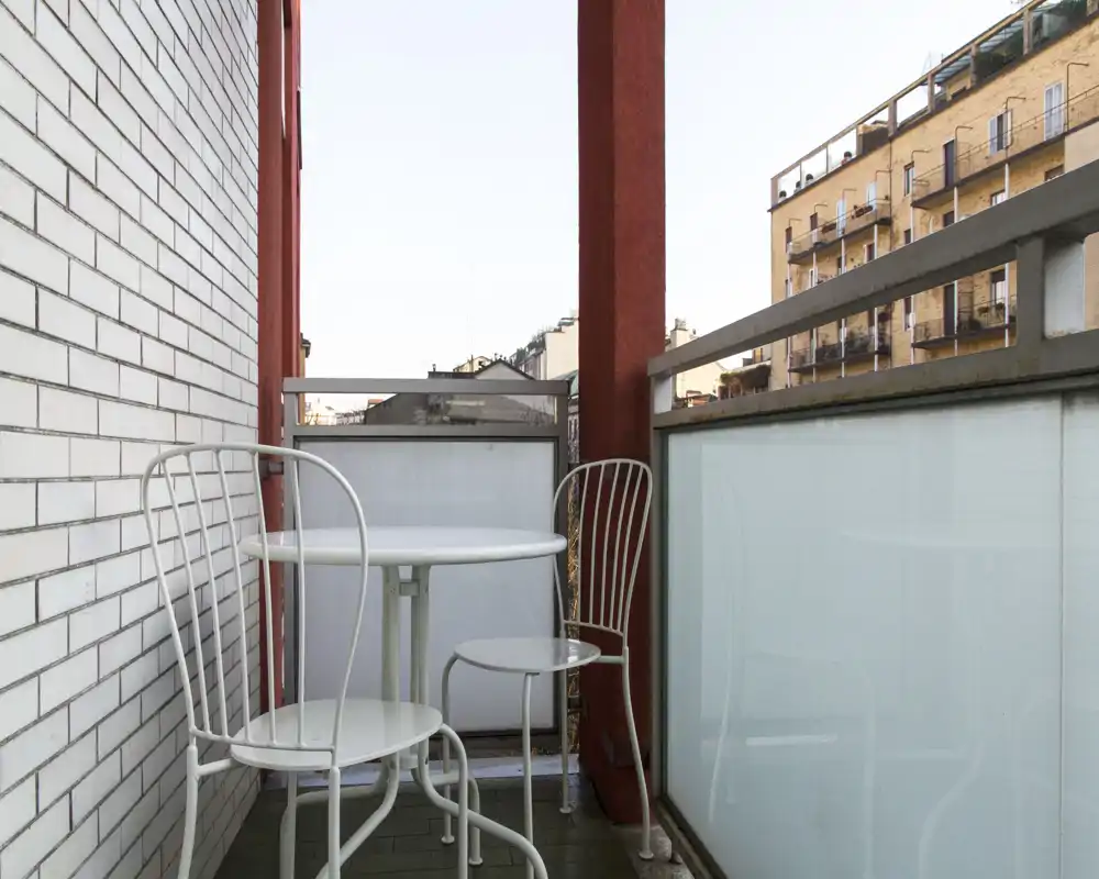 Balconcino cittadino compatto con piccolo tavolo rotondo e due sedie in metallo, parapetto in vetro satinato e vista sugli edifici vicini—perfetto per il caffè del mattino.