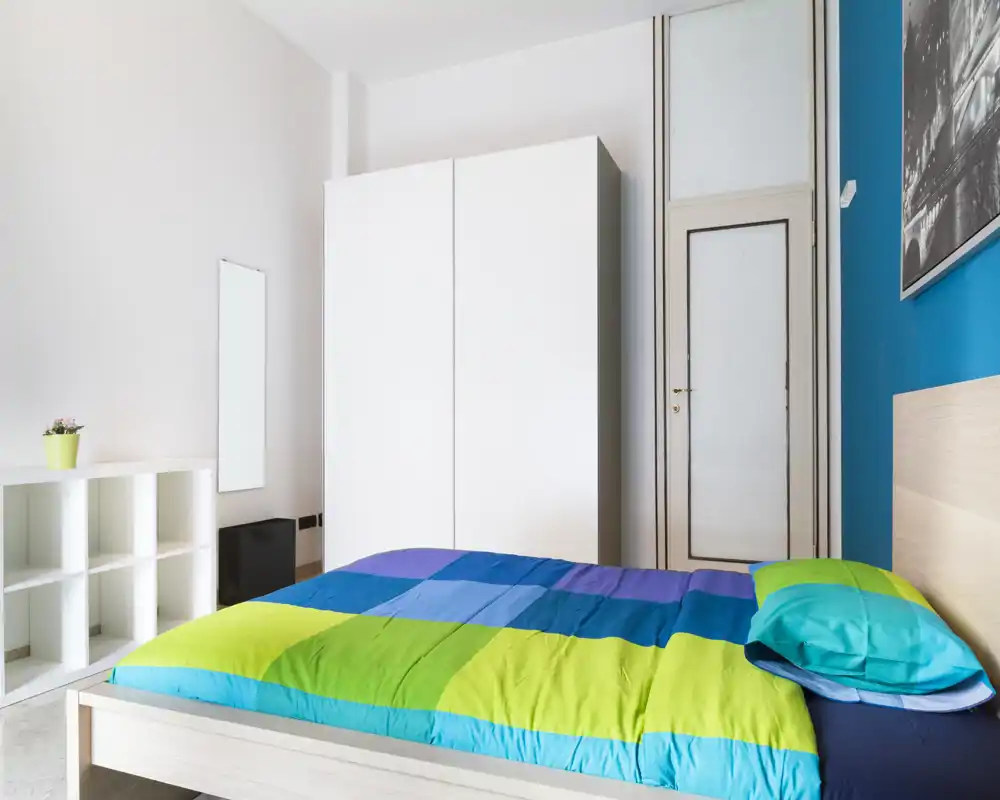 Camera da letto luminosa e moderna con letto matrimoniale colorato, armadio bianco e mensole minimaliste — uno spazio fresco e ben illuminato ideale per soggiorni brevi.