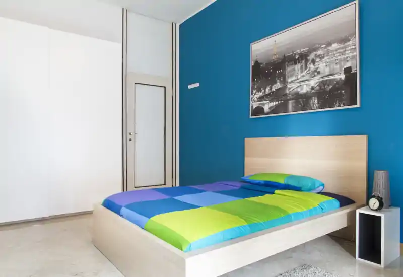 Chambre moderne et lumineuse avec lit plateforme en bois clair, couette colorée et mur d'accent bleu. Lignes épurées, bonne luminosité et décoration minimale la rendent accueillante et facile à mettre en valeur.