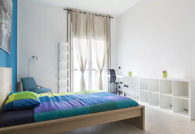 Chambre lumineuse et moderne avec un lit double en bois aux couvertures colorées, un fauteuil, un bureau et des étagères — idéale pour un séjour confortable avec beaucoup de lumière naturelle.