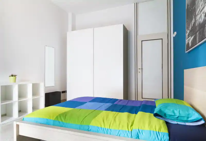 Chambre moderne et lumineuse avec un lit double coloré, une armoire blanche et des étagères minimalistes — un espace frais et bien éclairé, idéal pour de courts séjours.