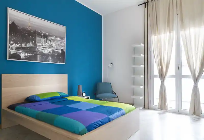 Chambre moderne et lumineuse avec un lit sur plate‑forme en bois et une literie colorée sur un mur bleu profond ; grandes fenêtres avec rideaux clairs apportent une belle lumière naturelle.