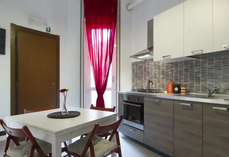 Cucina moderna con forno integrato, piano cottura a gas e lavello in acciaio accanto a un tavolo da pranzo per quattro con vaso decorativo; grande finestra con tenda rossa che illumina e scalda l'ambiente.
