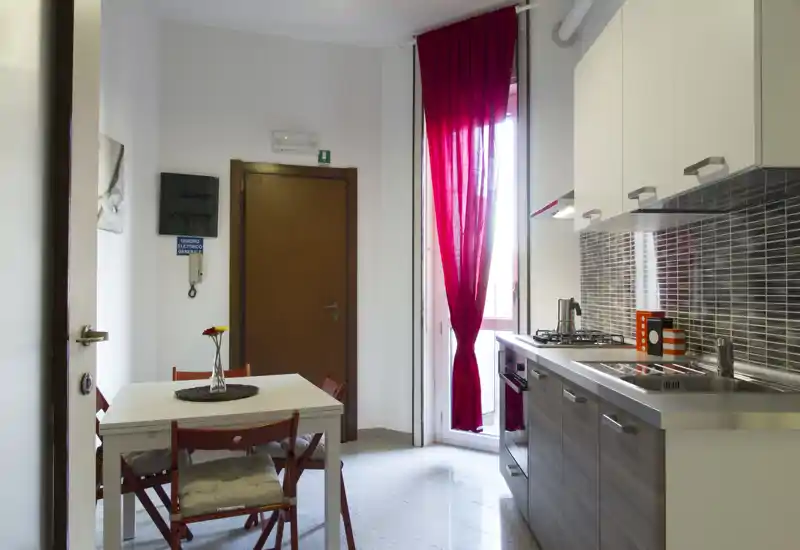 Cucina open-space compatta con pensili moderni, lavello in acciaio e un piccolo tavolo da pranzo per quattro persone. Luce naturale dall'alto della finestra e tocchi rossi che rendono l'ambiente accogliente.