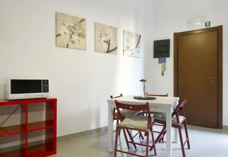 Piccola zona pranzo ordinata con tavolo bianco, sedie pieghevoli rosse e quadri alle pareti; luminosa e accogliente per un appartamento compatto.