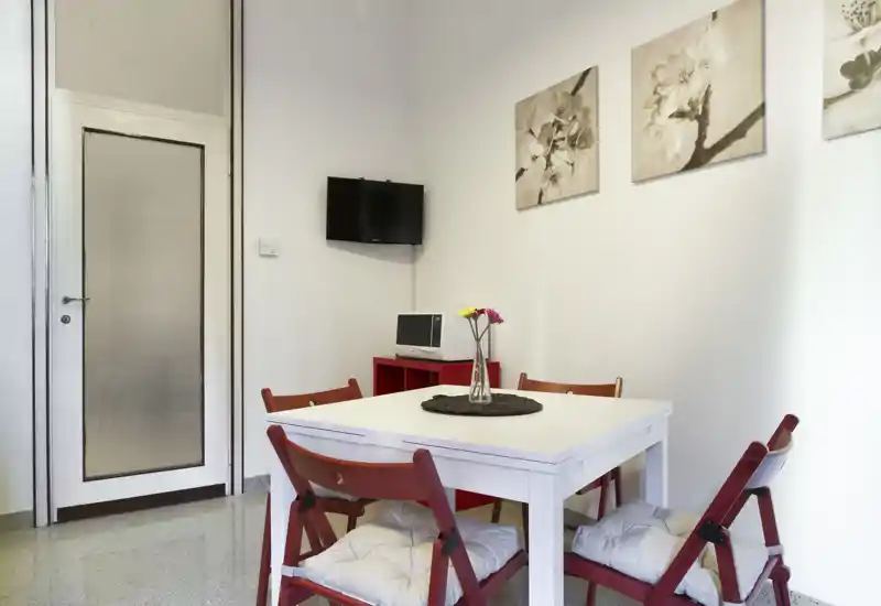 Zona pranzo luminosa e ben arredata con tavolo bianco, sedie pieghevoli rosse con cuscini e quadri decorativi; compatta e accogliente per i pasti.