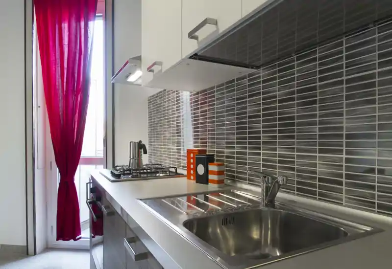 Cucina moderna e compatta con lavello in acciaio inox, piano cottura a gas e piastrelle lucide sul muro; una tenda rossa e piccoli accessori donano colore.