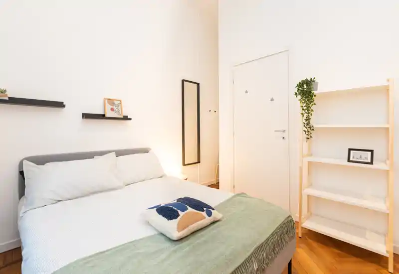 Camera da letto luminosa e moderna con letto matrimoniale ben rifatto, plaid verde e cuscino decorativo, pavimento in legno e scaffalatura essenziale — perfetta per un annuncio accogliente.