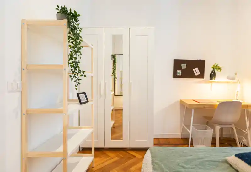 Camera da letto luminosa e minimal con angolo del letto visibile, armadio bianco con anta specchiata, scaffale in legno e una piccola scrivania — spazio pulito e ben illuminato, ideale per affitti brevi.
