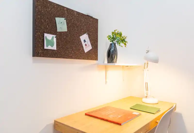 Angolo scrivania luminoso e ordinato con piano in legno, lampada da lavoro, bacheca in sughero e vaso decorativo — perfetto per uno studio o angolo lavoro.
