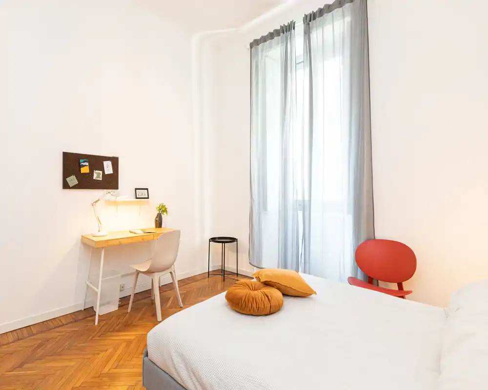 Bright, minimalist bedroom with a comfortable double bed, warm parquet flooring, a small desk and seating — ideal for a single traveler or couple.