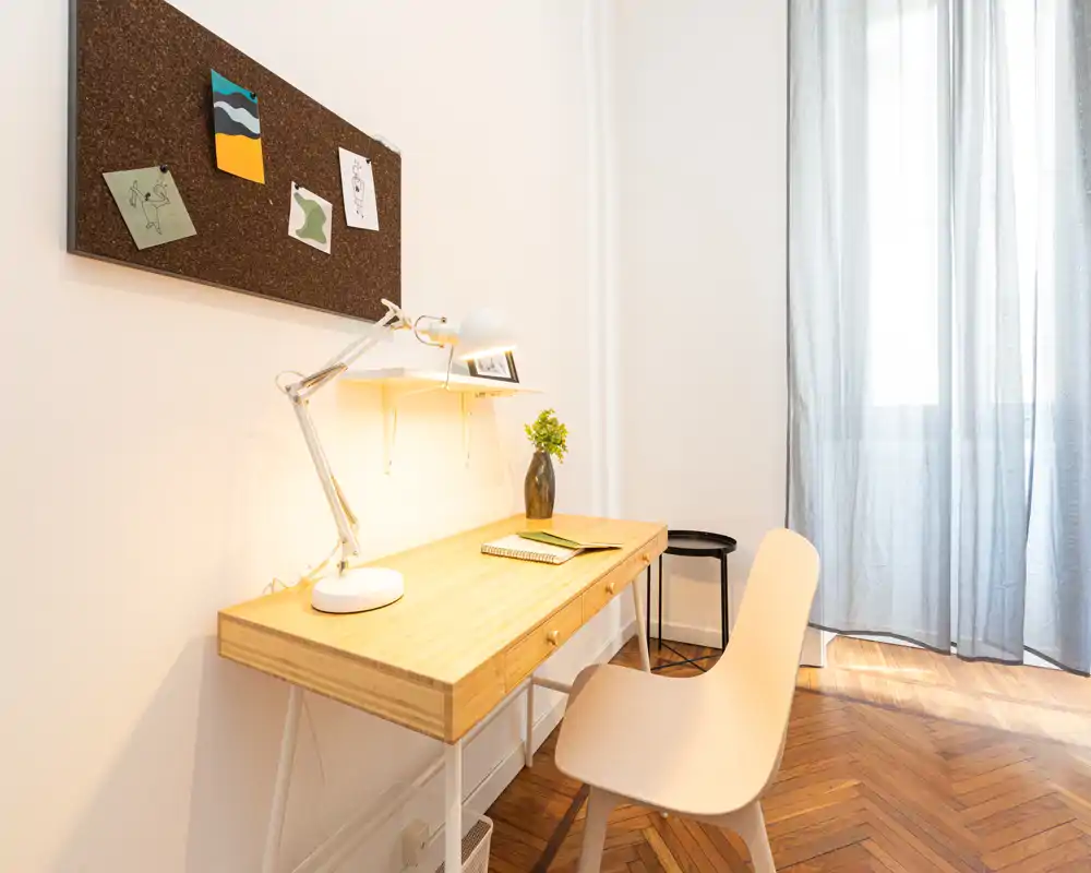 Bright, minimalist workspace with a wooden desk, task lamp, pinboard and a nearby window providing natural light—ideal for focused work or study.