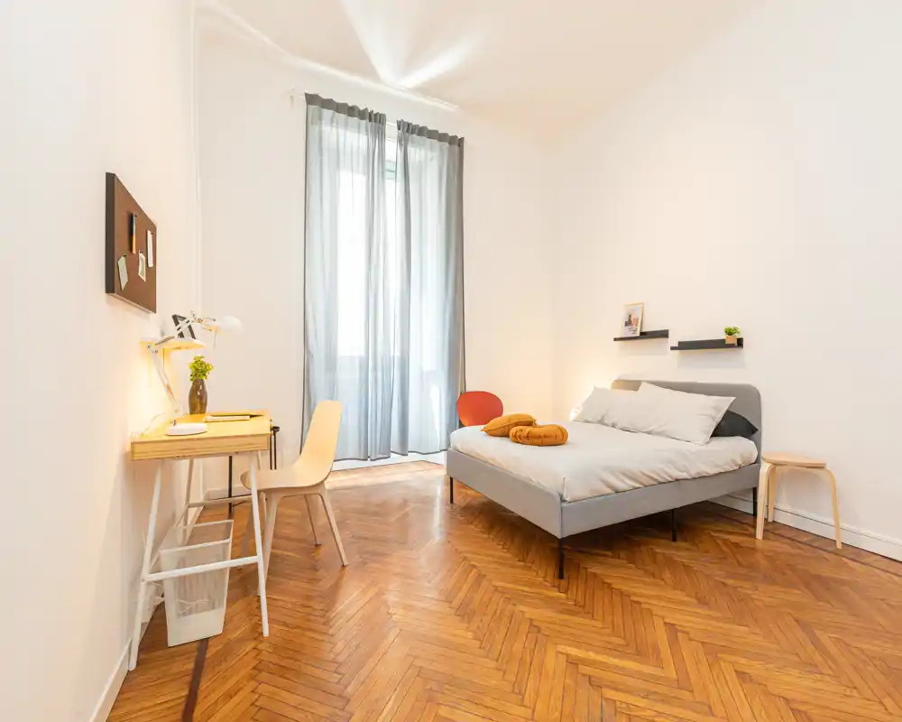Bright, minimal bedroom with a grey upholstered double bed, soft linens and warm herringbone parquet. A small desk sits by the window, creating a practical study corner with abundant natural light.