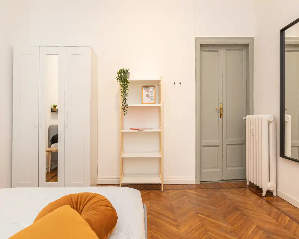 Bright, minimalist bedroom with warm parquet flooring, a visible bed with cozy cushions, a wardrobe and shelving — calm, well-lit space ideal for rental photos.