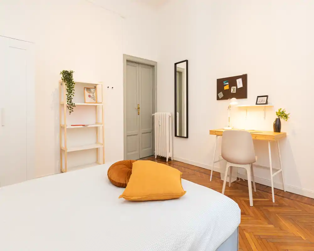 Bright, minimal bedroom with a large bed in the foreground, warm mustard cushions, a small wooden desk with lamp, and a full-length mirror — cozy and neatly styled for rental photos.