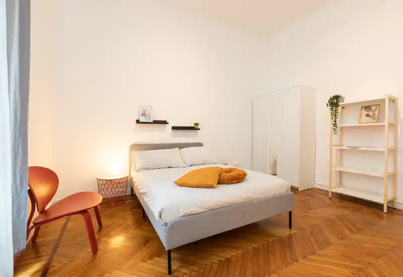 Bright, modern bedroom with a neatly made double bed, warm decorative cushions, a bedside lamp and minimalist storage. Hardwood herringbone floor and soft neutral walls create a calm, welcoming feel.
