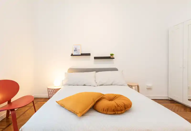 Bright, modern bedroom with a neatly made double bed, warm cushions and simple shelving above the headboard; minimal, cozy styling and wooden parquet flooring.