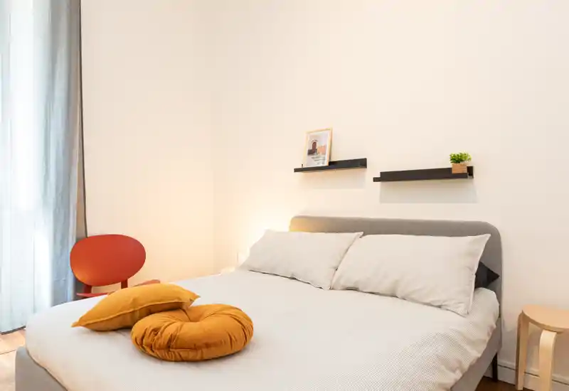 Bright, neatly styled bedroom with a comfortable double bed, soft neutral linens and mustard accent cushions. Large window with sheer curtains provides natural light; minimalist shelves and a chair complete the cozy layout.
