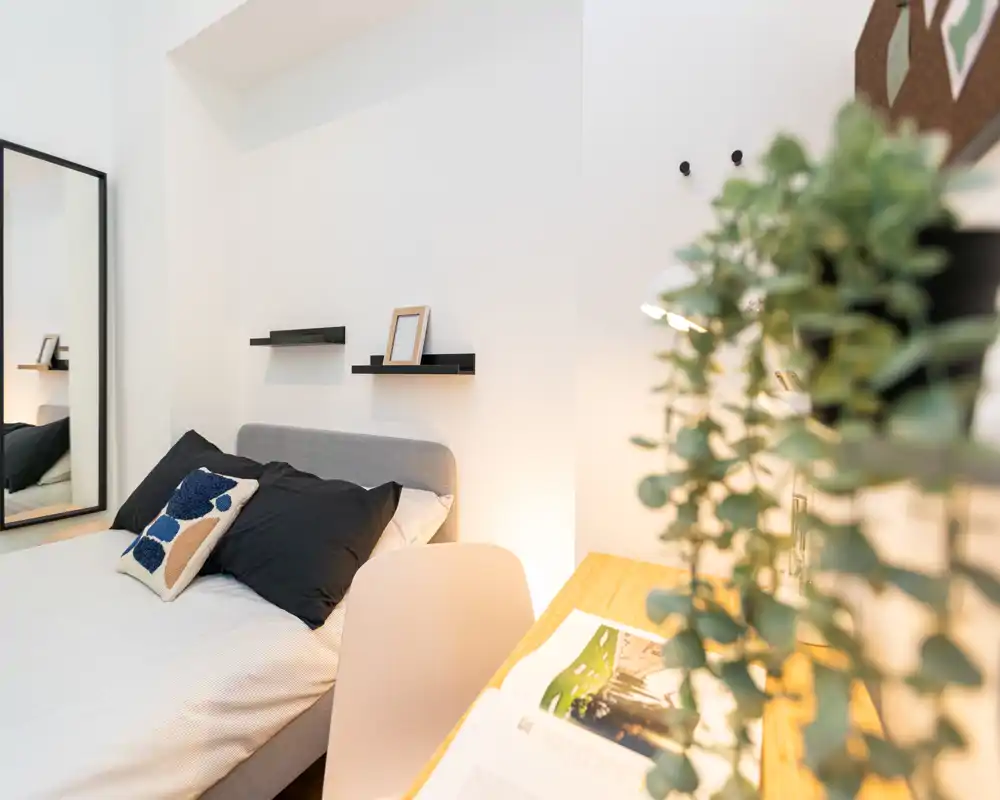 Camera da letto moderna e luminosa con un comodo letto matrimoniale, cuscini decorativi, mensole a parete e un'area scrivania — stile minimal e pulito, perfetto per annunci.