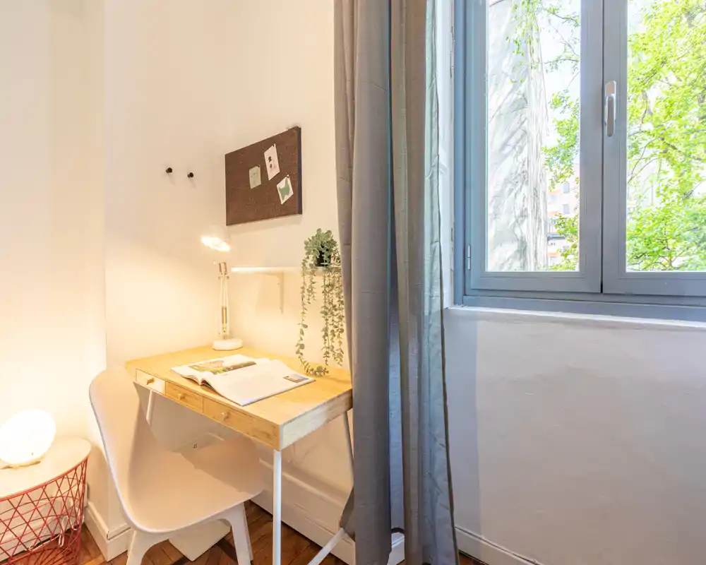 Angolo studio luminoso e minimal con scrivania in legno, sedia, lampada da tavolo e una finestra che offre luce naturale—ideale per lavoro o studio.