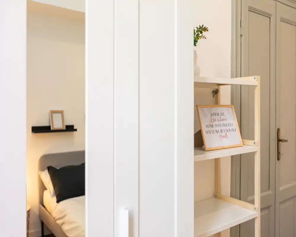 Vista parziale di una camera da letto dietro un divisorio bianco: letto ordinato con cuscini e testiera morbida, lampada da comodino calda e mensole decorative minimal.