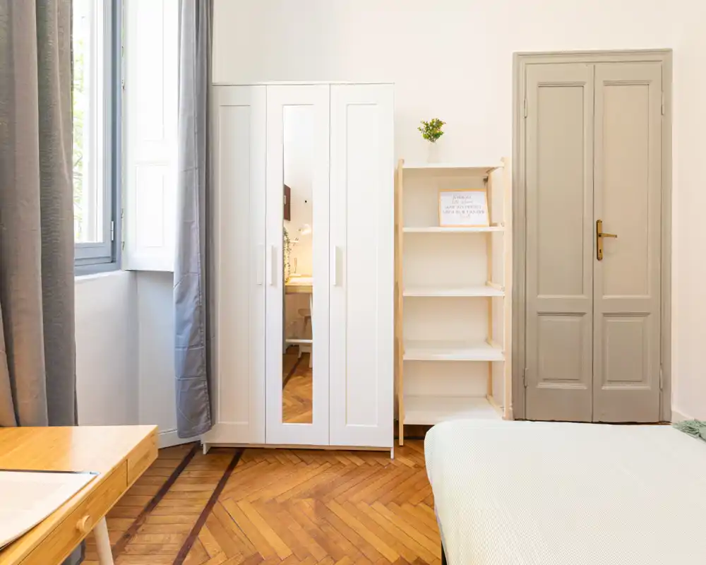Camera da letto luminosa e minimal con vista parziale del letto, armadio bianco con anta a specchio, scaffalatura aperta e ampia finestra che lascia entrare luce naturale.