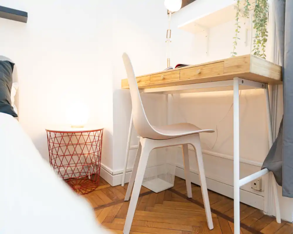 Un angolo di camera da letto compatto con lampada da comodino, scrivania moderna e sedia accanto al letto; luminoso, minimal e funzionale per lavorare o leggere.