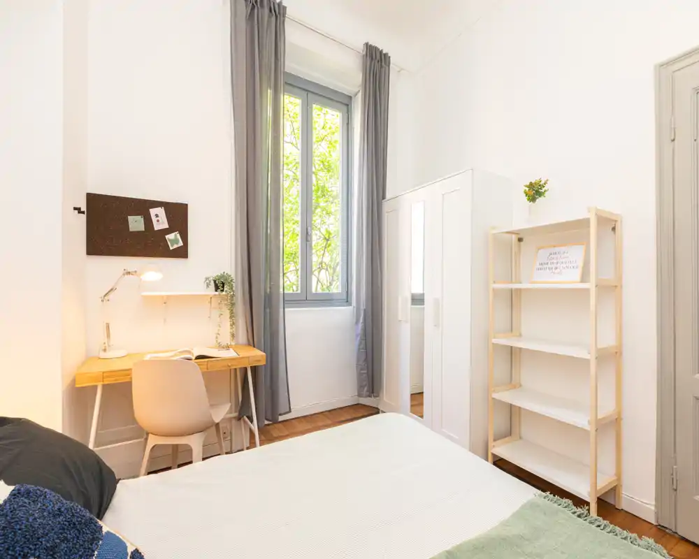 Camera da letto luminosa e ariosa con letto ben rifatto, scrivania in legno vicino alla finestra e armadio — perfetta per un soggiorno comodo da single.