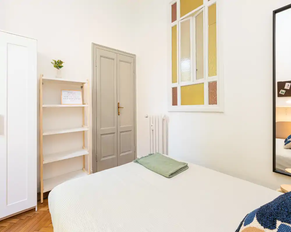 Camera da letto luminosa e ordinata con letto matrimoniale, biancheria dai toni morbidi e arredi minimal; pavimento in legno e specchio decorativo creano un’atmosfera accogliente.